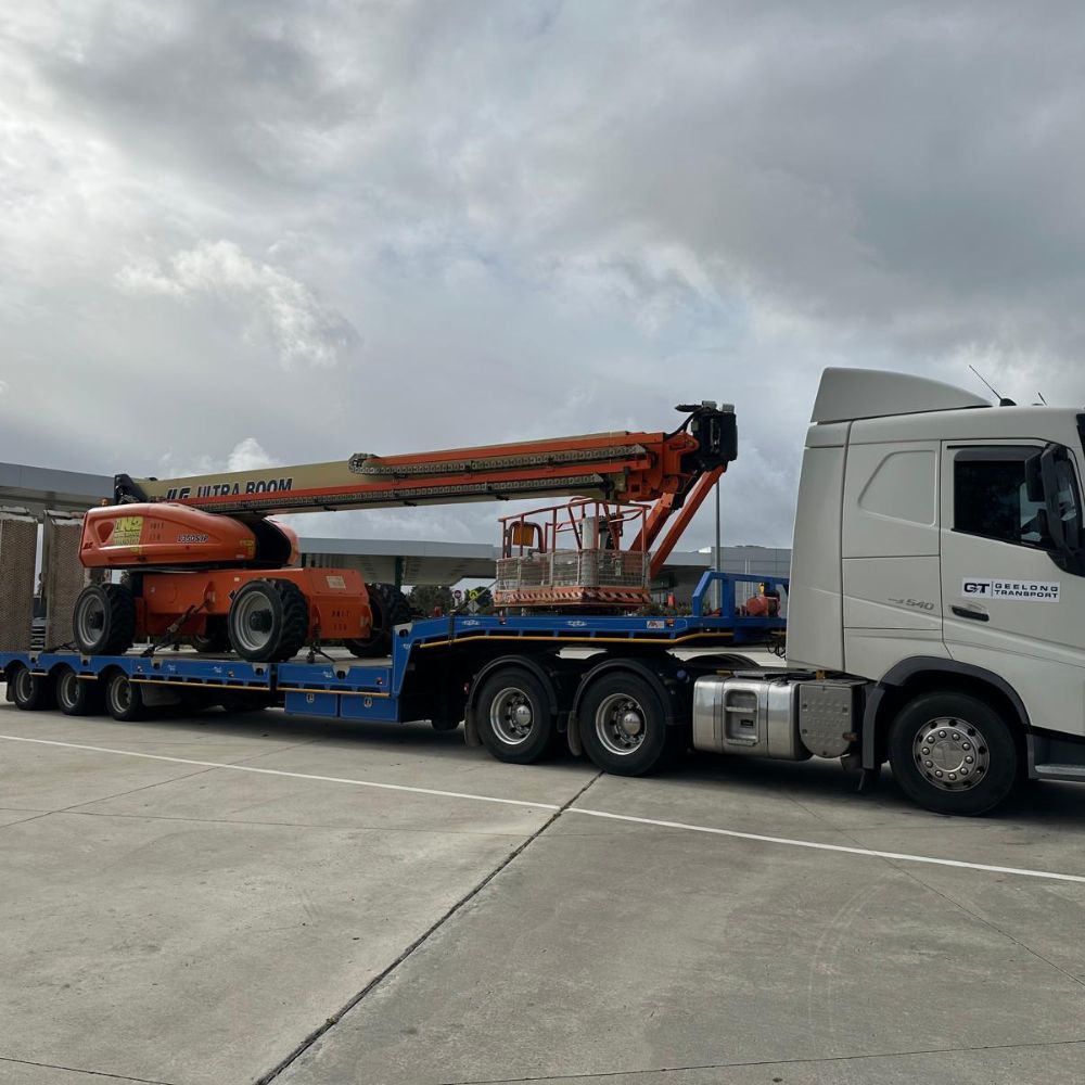 A truck carries an orange cherry picker on a flatbed under cloudy skies.